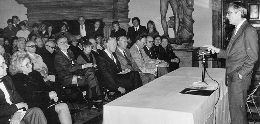 A man speaking at a table and microphone to seated audience.
