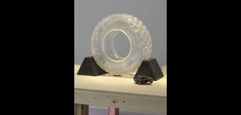 Clear mold of a tire held upright by two dark pyramid shaped blocks on a light wooden table with a gray background.