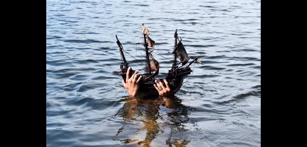 Two hands holding a small old-fashioned model ship (about one and a half feet long) above water in the daytime.