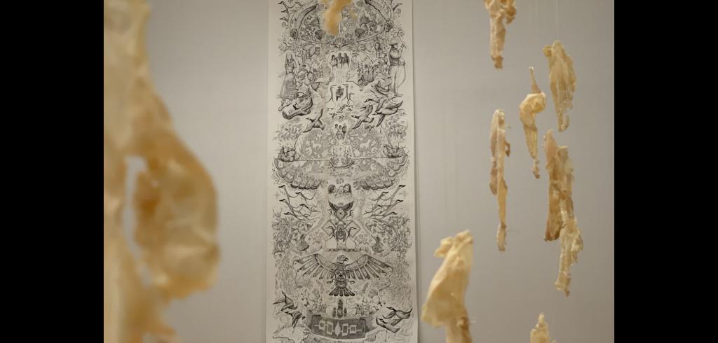 Abstract cream colored figures hanging in front of a black and white print of different scenes in nature.