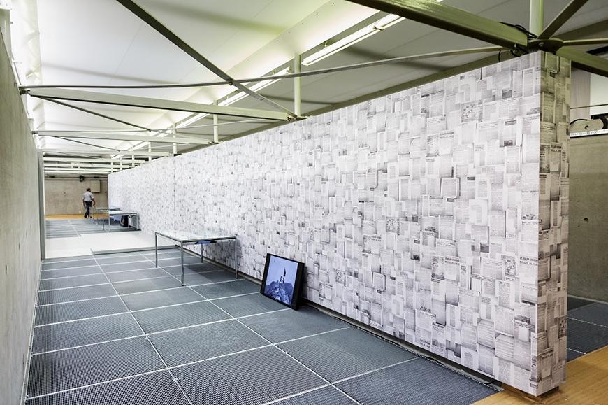 gallery wall papered with newspaper clippings with two tables and an LCD screen propped against it