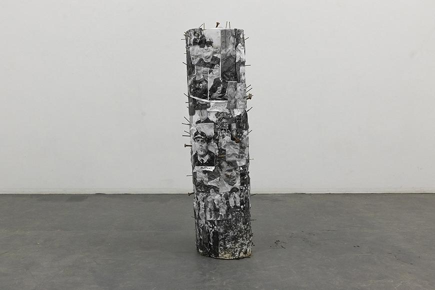 A wooden column covered in black and white photographs and protruding nails.