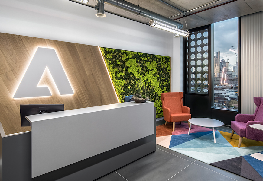 Office entrance with a large desk and computers. Along side is a colorful sitting arear by a window with a tabkle and 2 chairs. 