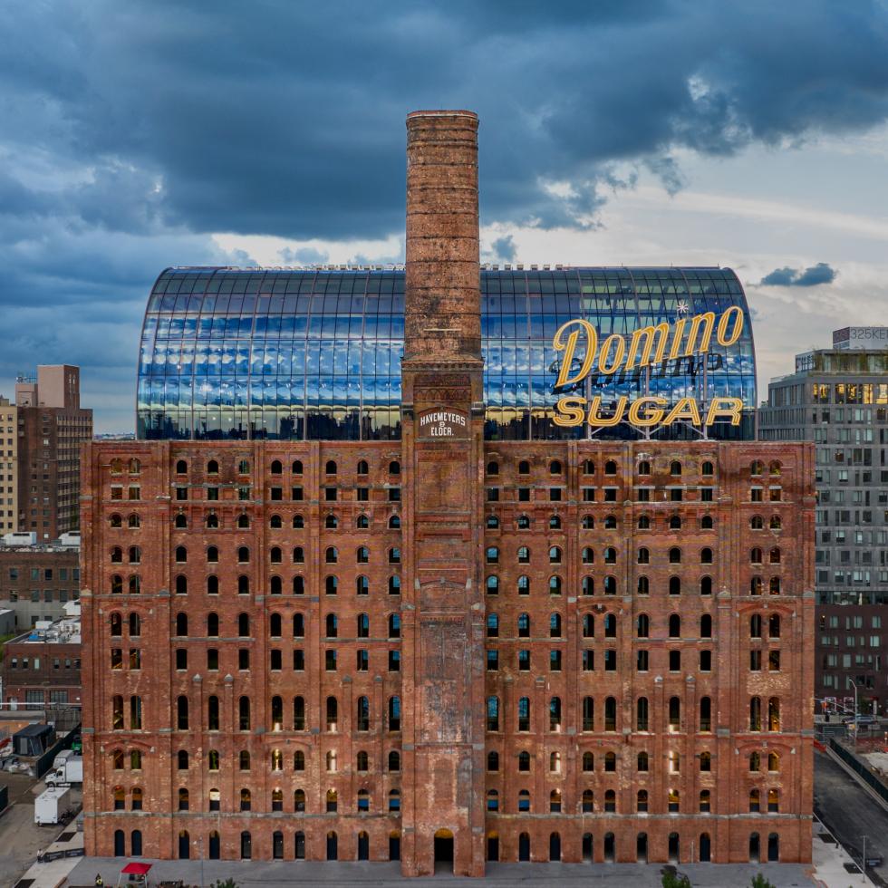 An aerial view of the historic Domino Sugar Refinery in Brooklyn, New York. The brick building is topped with a modern glass structure and a large 