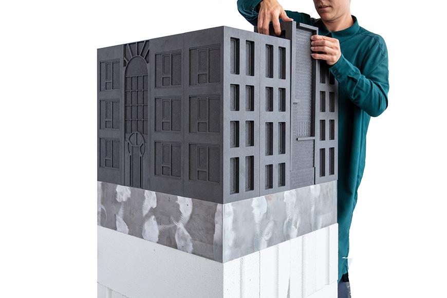 A model of a building which has been rendered entirely in grey material, and placed within a gallery space. The model is elevated and placed on a white and grey podium. 