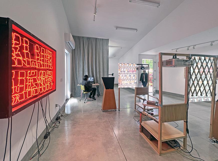 A large white room with a neon red sign on the left wall, a wooden sculpture in the middle of the floor, and a person sitting at a desk in the background.
