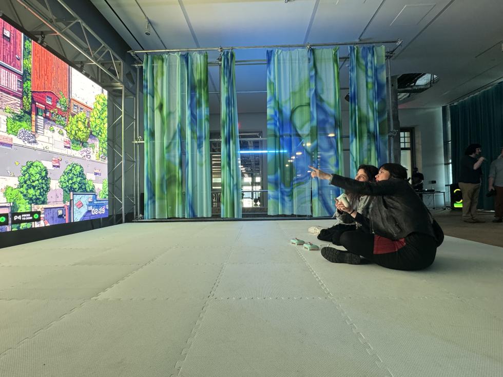 Side-view of two young people talking, holding consoles, and pointing at a large screen showcasing a videogame.