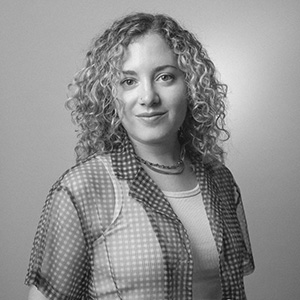 A person with light skin and curly blonde hair, wearing a sheer polka dot blouse over a white tank top.
